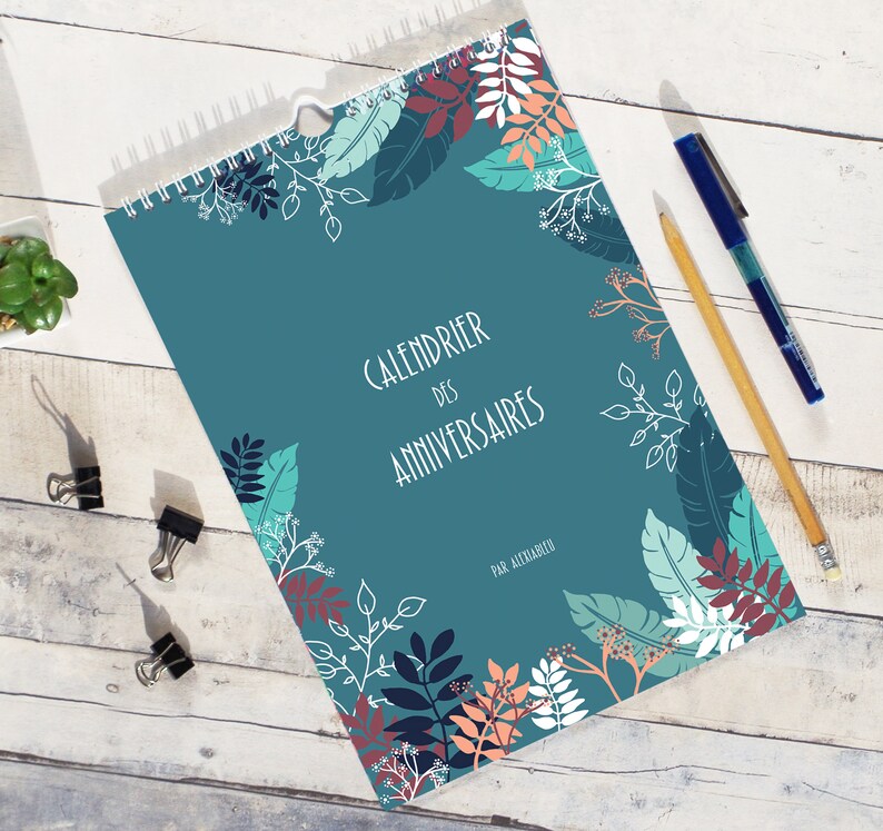 May include: A spiral-bound birthday calendar with a teal cover featuring a botanical pattern of leaves and branches in shades of dark blue, light blue, white, and peach. The text reads "CALENDRIER DES ANNIVERSAIRES" and "PAR ALEXIABLEU".