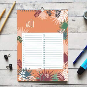May include: A wall calendar page for the month of August, labeled with the text AO&Ucirc;T, featuring a peach-colored background decorated with botanical illustrations of leaves and flowers in shades of green, white, and dark purple, with a white grid.