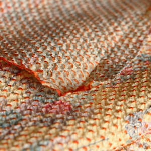 May include: Close-up of a textured fabric with a floral pattern. The fabric is a mix of cream, orange, and pastel colours, with visible stitching. The stitching is in orange thread, creating a dotted pattern.