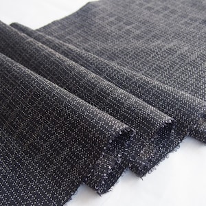 May include: A length of dark charcoal grey fabric featuring a subtle, repeating grid pattern created by tiny white flecks. The textile has a textured, woven appearance with visible frayed edges along the sides and soft, cascading folds.