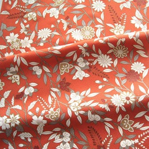 May include: Close-up of a fabric with a floral pattern. The fabric is a vibrant orange-red color, with a repeating design of white, gold, and burgundy flowers and gray leaves. The fabric has a subtle diagonal texture.