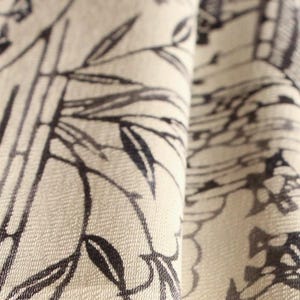 Può includere: Primo piano di un tessuto piegato con un motivo toile de Jouy in bianco e nero. Il design presenta steli di bambù, foglie ed elementi architettonici. Il tessuto ha un aspetto strutturato e una palette di colori neutri.