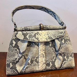 Könnte beinhalten: Eine Vintage-Handtasche mit Schlangenmuster in Grau-, Schwarz- und Cremetönen. Die Tasche hat einen geschwungenen Klappenverschluss, einen oberen Griff und einen Metallverschluss. Das Gesamtdesign ist strukturiert und klassisch.