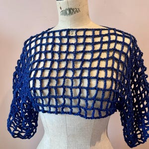 May include: A dark blue, open-weave crochet top with short sleeves is displayed on a mannequin. The top has a square grid pattern and is made of a shiny, possibly metallic, yarn. The mannequin is a neutral colour, and the background is a soft pink.