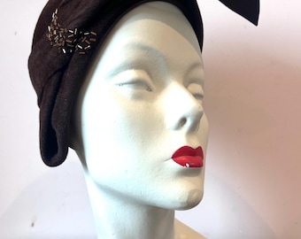 May include: A dark brown felt hat featuring a structured, asymmetrical brim that sweeps upward on one side. The hat is adorned with a small cluster of metallic, cylindrical beads on the side, adding a subtle texture to the smooth, dark fabric.