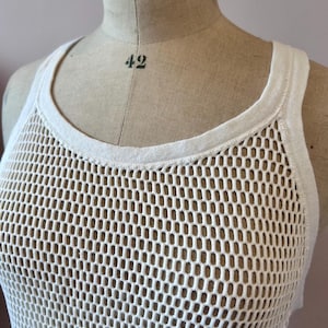 May include: Off-white, open-weave tank top. The sleeveless top features a round neckline and a honeycomb-like mesh design. The garment is displayed on a mannequin with the word "STOCKMAN" and the number "42" visible.
