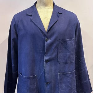 May include: A navy blue work jacket with three front pockets and a three-button closure. The jacket has a notched lapel collar and is displayed on a mannequin. The jacket is made of a durable fabric.