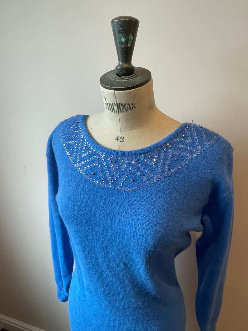 May include: A light blue sweater with a sequined neckline. The sequins are iridescent and create a geometric pattern.