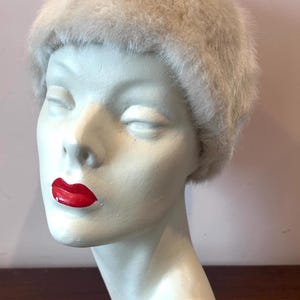 Peut inclure: Un chapeau de fourrure beige clair de style vintage est posé sur une tête de mannequin aux yeux fermés et aux lèvres rouges. Le chapeau a une forme arrondie et un aspect doux et texturé. Le mannequin est peint en blanc, sur fond rose.