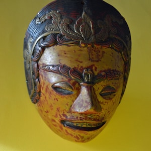 May include: A wooden mask with a human face, painted in gold, red, and black. The mask has intricate details, including a crown of leaves and a stylised nose.