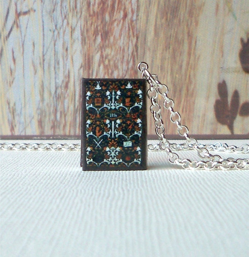 May include: A silver chain necklace with a small pendant shaped like a book. The book cover is brown with a black and white patterned design.