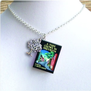 May include: A silver chain necklace with a charm of a book titled "A Tree Grows in Brooklyn" by Betty Smith and a silver tree charm.