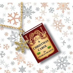 May include: A gold-colored necklace with a snowflake charm and a miniature book pendant. The book cover is burgundy with gold lettering that reads "A Christmas Carol" by Charles Dickens. The background features snowflakes.