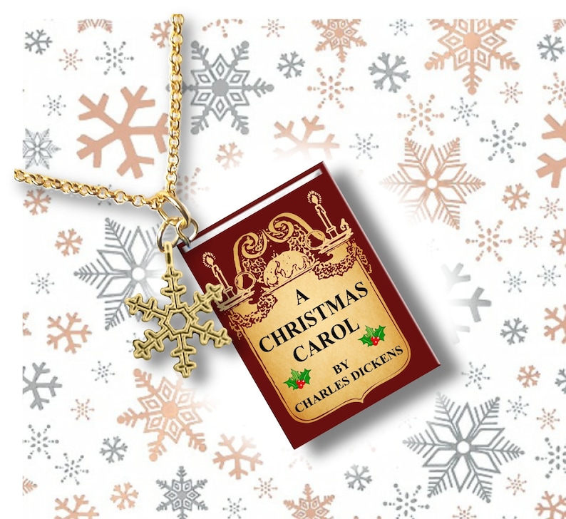 May include: A gold-colored necklace with a snowflake charm and a miniature book pendant. The book cover is burgundy with gold lettering that reads "A Christmas Carol" by Charles Dickens. The background features snowflakes.