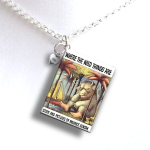 May include: A silver necklace with a charm featuring the cover art of the children's book "Where the Wild Things Are" by Maurice Sendak. The charm is a rectangle with a colorful illustration of a wild thing sitting on a red boat in a jungle setting.