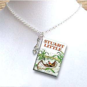 May include: A silver chain necklace with a charm of a mouse in a canoe. The charm is attached to a small, rectangular pendant with the words "Stuart Little" printed on it. The pendant is a replica of the book cover.