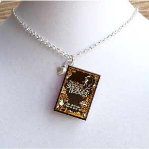 May include: A silver chain necklace with a charm of a book cover. The book cover is brown with gold lettering and a silhouette of a man. The text on the book cover reads "The Complete Sherlock Holmes" and "Sir Arthur Conan Doyle". A small silver heart charm hangs from the chain above the book charm.
