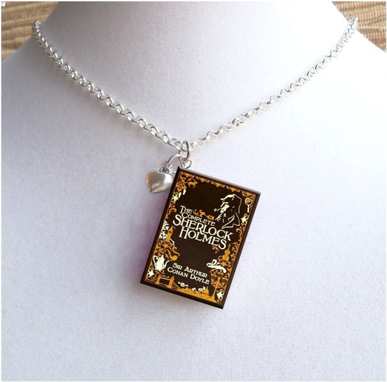 May include: A silver chain necklace with a charm of a book cover. The book cover is brown with gold lettering and a silhouette of a man. The text on the book cover reads "The Complete Sherlock Holmes" and "Sir Arthur Conan Doyle". A small silver heart charm hangs from the chain above the book charm.