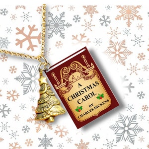 May include: Gold-colored Christmas tree pendant necklace with a chain. The pendant is next to a small book with the title "A Christmas Carol" by Charles Dickens. The background has snowflakes.