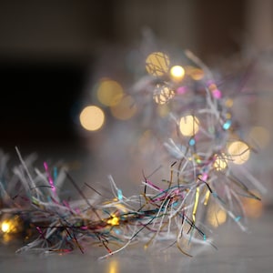 Op de afbeelding: Een witte, glinsterende guirlande met meerkleurige strengen tinsel en kleine, warmwitte lampjes.