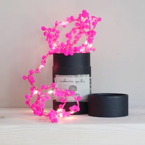 May include: A string of bright pink pom-pom lights with small white lights, partially displayed from a black cylindrical box. The box has a label that reads "melanie porter". The lights are on a white surface.