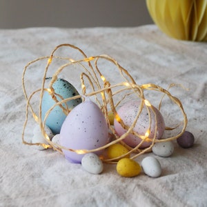 May include: An Easter-themed arrangement featuring speckled pastel eggs in blue, pink, and lavender, accented with small white and yellow speckled eggs. Twinkling fairy lights are woven around the eggs, creating a festive display. A yellow paper honeycomb ball is in the background.