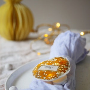 Peut inclure: Une carte de table en tissu jaune avec le nom "Tilly" écrit dessus. La carte de table est éclairée de l'intérieur et est posée sur une serviette bleue.