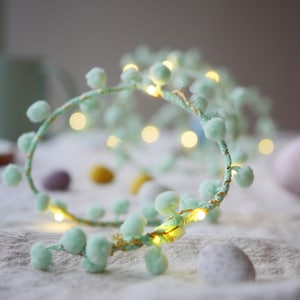 May include: A string of fairy lights with small, light green pom-poms. The lights are wrapped around a gold wire and are lit, casting a warm glow.
