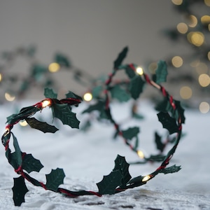 May include: A string of fairy lights with green holly leaves and red wire. The lights are strung together in a circle and are lit up. The lights are on a white surface.