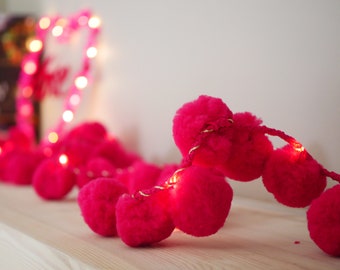 May include: A string of pink pom-pom lights with warm white LED lights. The lights are strung together with a gold wire.