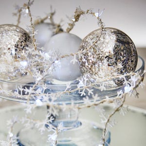 May include: A festive arrangement of silver and white Christmas ornaments on a clear glass cake stand. A garland of white snowflake cutouts with gold accents drapes around the ornaments, creating a winter holiday theme.