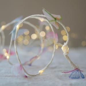 May include: A string of fairy lights with small, delicate butterfly decorations in shades of pink, blue, and green. The lights are on a white wire, and the butterflies appear to be made of translucent paper. The background is softly blurred.