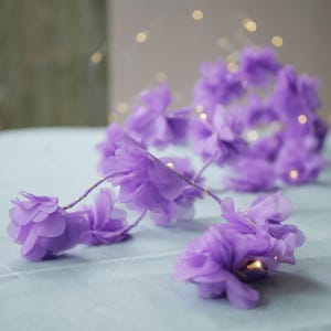 Op de afbeelding: Een slinger van paarse stoffen bloemen met kleine witte lampjes die erdoorheen geweven zijn. De bloemen zijn in een cascade-achtige opstelling gerangschikt, wat een delicate en romantische uitstraling creëert.