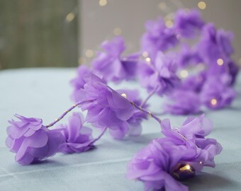 May include: A string of purple fabric flowers with small white lights woven through them. The flowers are arranged in a cascading pattern, creating a delicate and romantic look.