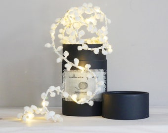 May include: White fairy lights with fluffy pom poms wrapped around a black tube with a label that says "melanie + me" and a small illustration of a ball of yarn.