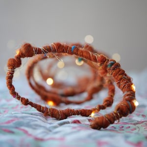 Puede incluir: Guirnalda decorativa artesanal de color marrón anaranjado con pequeñas luces LED blancas cálidas. La guirnalda está hecha de hilo y alambre envueltos, con detalles plateados y coloridos. Las luces están espaciadas uniformemente, creando un brillo suave.