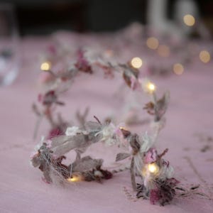 Peut inclure: Une guirlande lumineuse décorative composée de bandes de tissu entrelacées dans des tons rose poudré, crème et gris. La guirlande est ornée de textures textiles effilochées et de petites ampoules LED aux tons chauds diffusant une lueur douce.