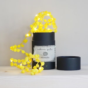 May include: Yellow pom pom string lights wrapped around a black tube with a white label that says "melanie porter" and a small illustration of a ball of yarn. The lights are battery operated and are perfect for adding a touch of whimsy to any space.