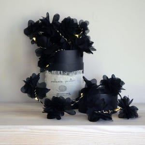 May include: Black floral string lights with a gold wire, displayed in and around a black round box. The lights are arranged on a white surface. The box has a white fabric with the text "melanie porter" on it.