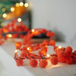 Puede incluir: Una guirnalda decorativa que presenta una serie de pompones de color naranja brillante unidos a un cordón oscuro trenzado, intercalados con pequeñas luces LED de color blanco cálido que emiten un suave resplandor a lo largo de la cuerda.