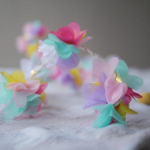 May include: A pastel-colored flower garland with a string of lights. The garland is made of multiple layers of tulle fabric in shades of pink, yellow, blue, and purple.