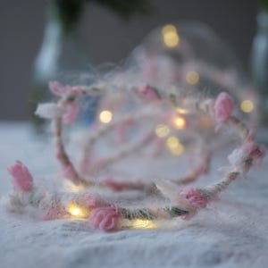 May include: A handmade floral crown with pink fabric flowers and white accents. The crown is wrapped in a light-colored yarn and features small, warm white LED lights. The crown is set against a soft, textured background.