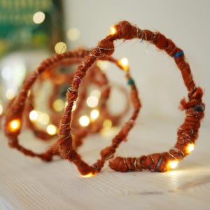 Op de afbeelding: Een decoratieve lichtsnoer met een flexibele draad omwikkeld met getextureerd, roestoranje garen. Het garen is geaccentueerd met dunne, zilverkleurige metaaldraad en kleine, kleurrijke vezels. Warmwitte LED-lampjes zijn erin verwerkt.