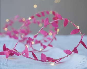 May include: A string of pink leaf-shaped fairy lights. The leaves are arranged along a thin, twisted wire, with small, warm white LED lights interspersed. The lights are on, creating a soft, ambient glow. The garland is on a white surface.