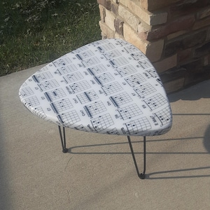May include: A triangular stool with a white top featuring a black printed pattern of various guitar chord diagrams and labels. The stool is supported by three thin, black metal hairpin legs that provide a stable base for the seat.