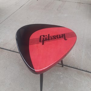 May include: A triangular side table with a red and black top, featuring the "Gibson" logo. The table has three black legs and a white trim. The table measures approximately 24 inches wide and 18 inches tall.