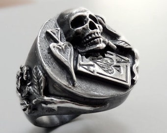 Puede incluir: Un anillo de plata con un diseño de calavera y huesos cruzados. La calavera está detallada y tiene una expresión amenazante. El anillo está engastado con una piedra negra y la banda está decorada con un diseño floral.