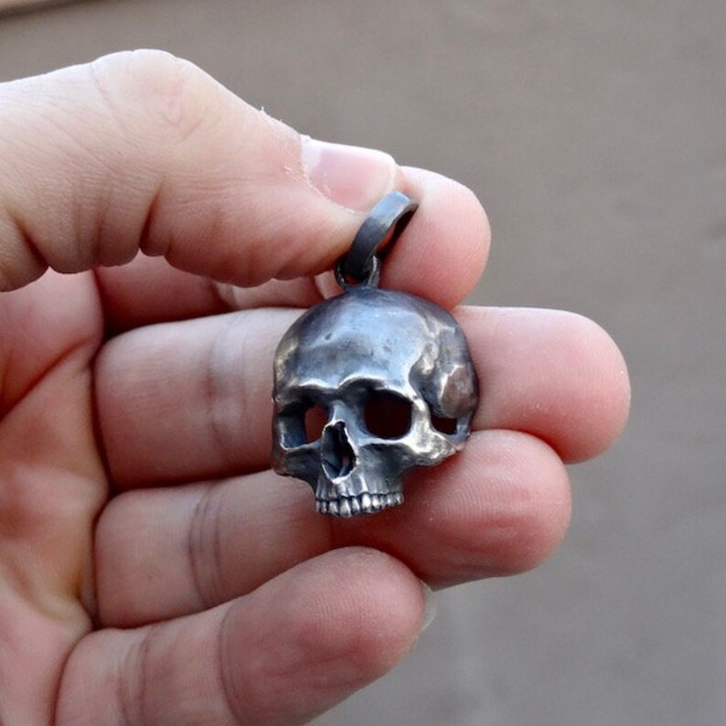 May include: A silver skull pendant with a detailed design. The pendant is hanging from a silver loop.