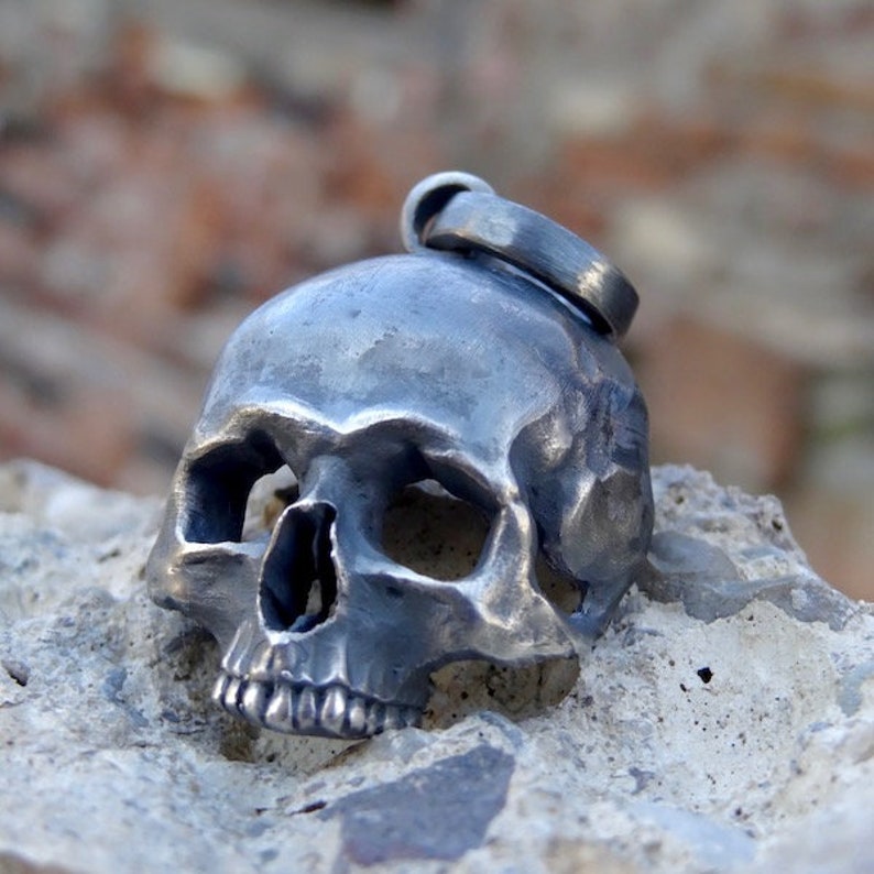 May include: A silver skull pendant with a detailed design. The skull has a textured surface and is slightly tilted to the side. The pendant has a loop at the top for attaching to a chain.