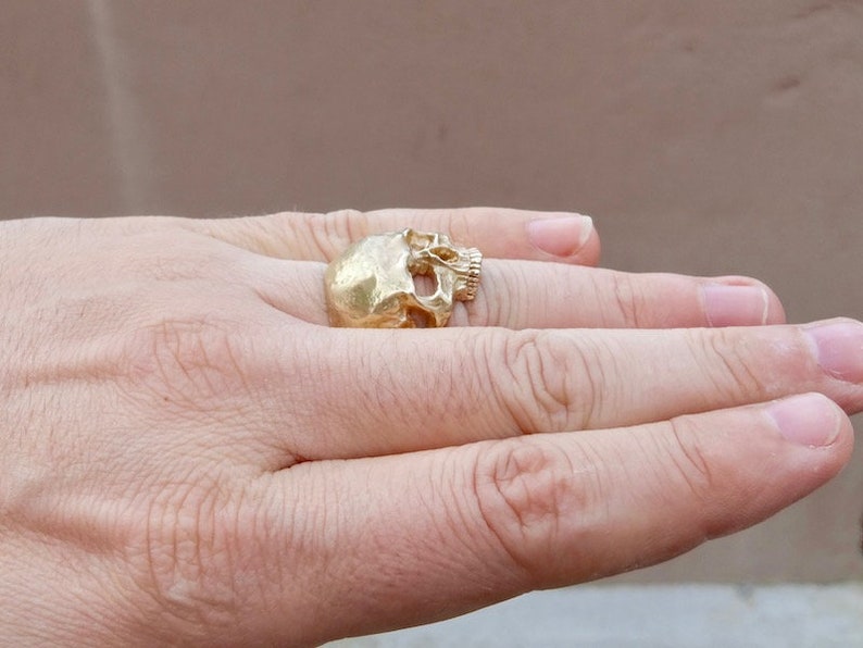 May include: A gold-colored skull ring worn on a person's finger.